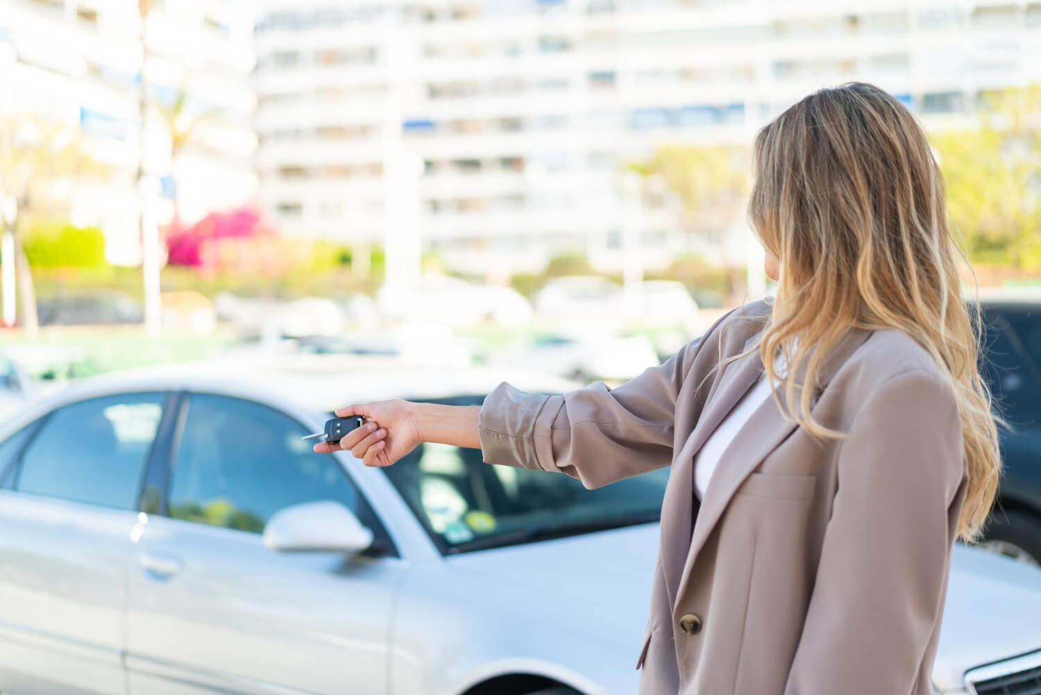Donna con capelli biondi in abbigliamento elegante punta il telecomando verso un'auto parcheggiata, concetto di noleggio auto a lungo termine o scelta del veicolo.